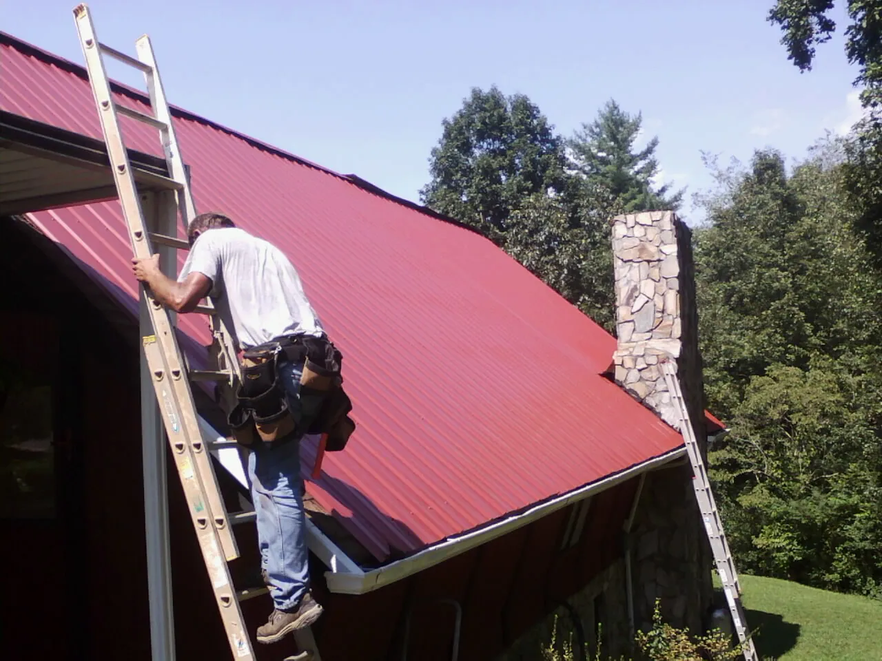 Roofer on a ladder beside a red metal roof and stone chimney — Soffit & Fascia Repair by Roofing Brothers in Pearland